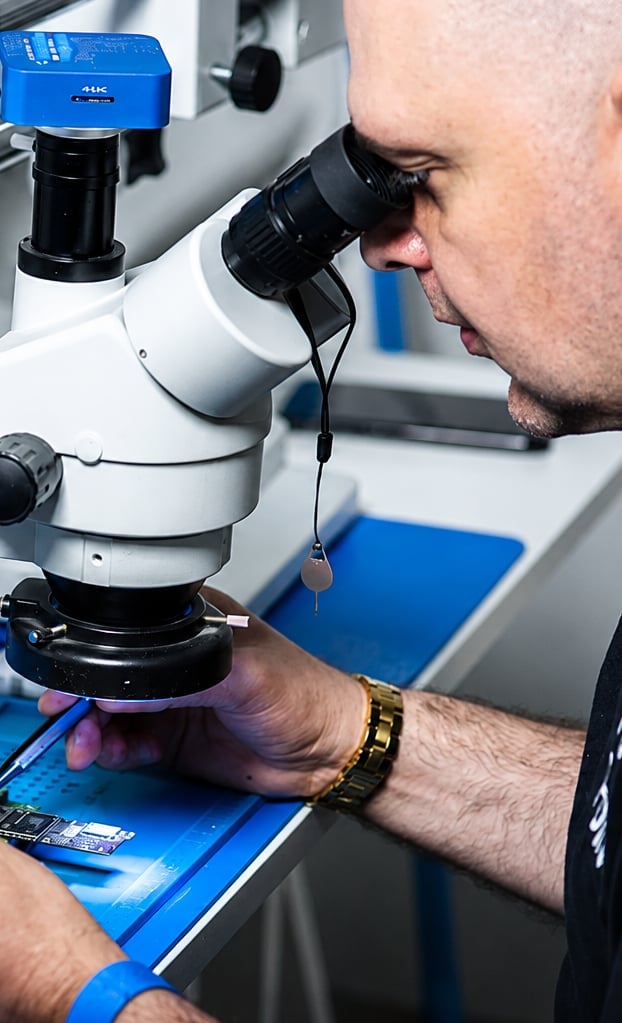 Technician examining a circuit board through a blue stereo microscope in a laboratory setting