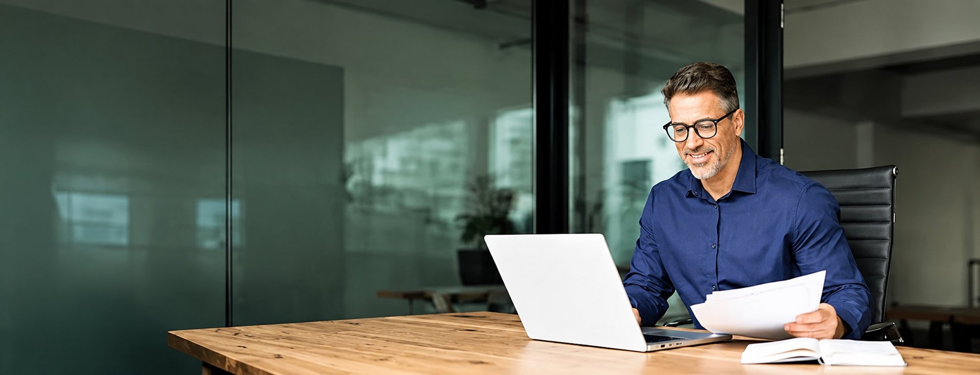 Man in blue shirt working at wooden desk with laptop and documents in modern office with glass walls