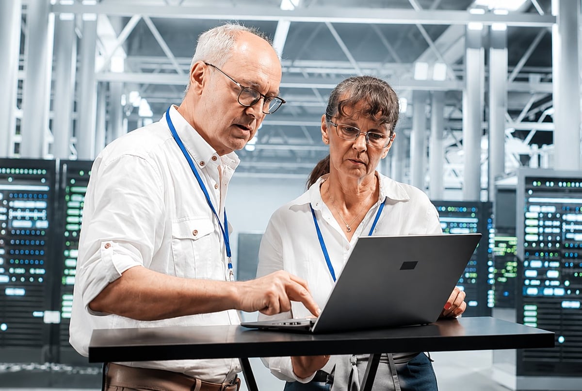 Two technicians in white shirts examine a laptop together in a data center control room with server equipment behind them
